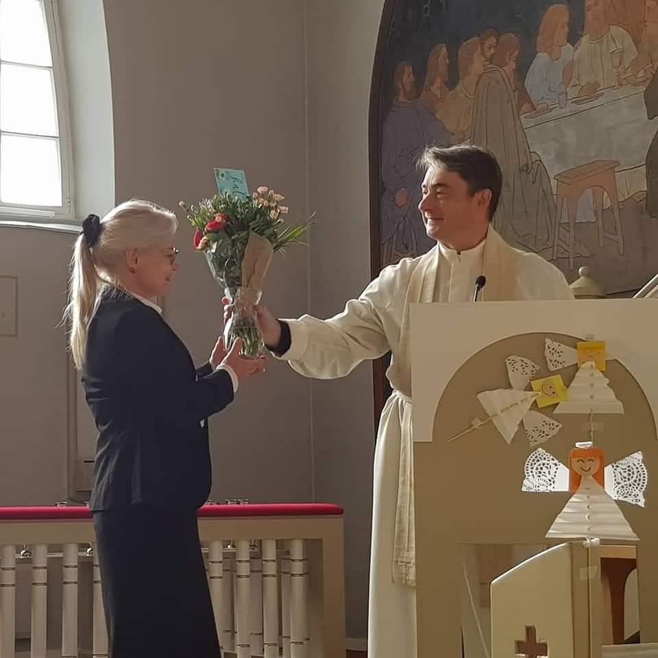 Kirkkoherra Timo Vainikainen ojentaa kukkia seurakuntamestari Virpi Voutilaiselle.kirkon alttarin edustalla.